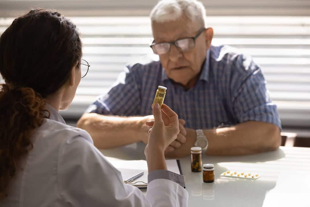 A female doctor in a white coat holds up a pill bottle to show an elderly male patient sitting across from her at a table with several medication bottles
