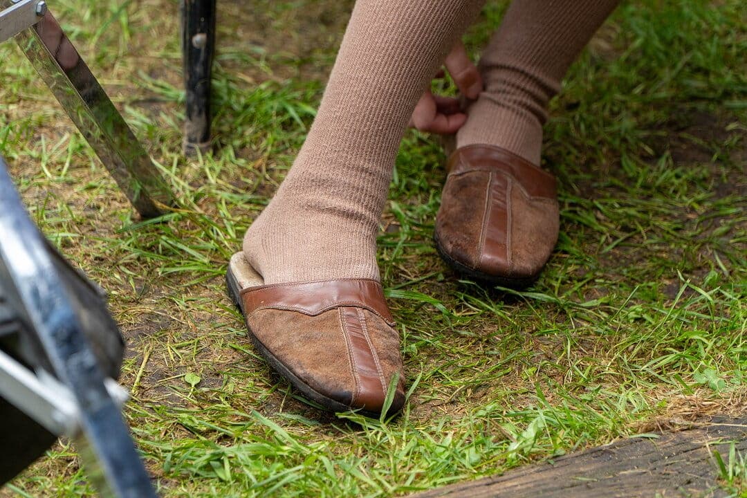 Gros plan sur les pieds d'une personne âgée chaussée de mocassins marron amples et de chaussettes beiges, à l'extérieur sur l'herbe, avec un déambulateur à proximité