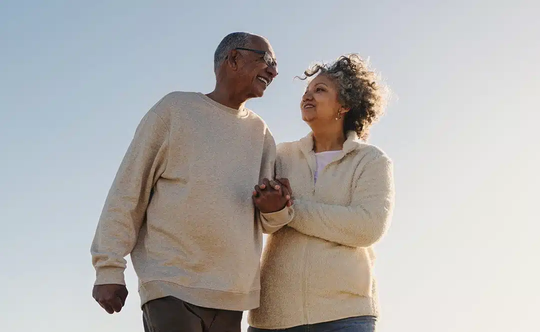 Un couple de personnes âgées, l'air heureux, se tient la main et s'échange des sourires tout en se promenant en plein air sous un ciel bleu limpide
