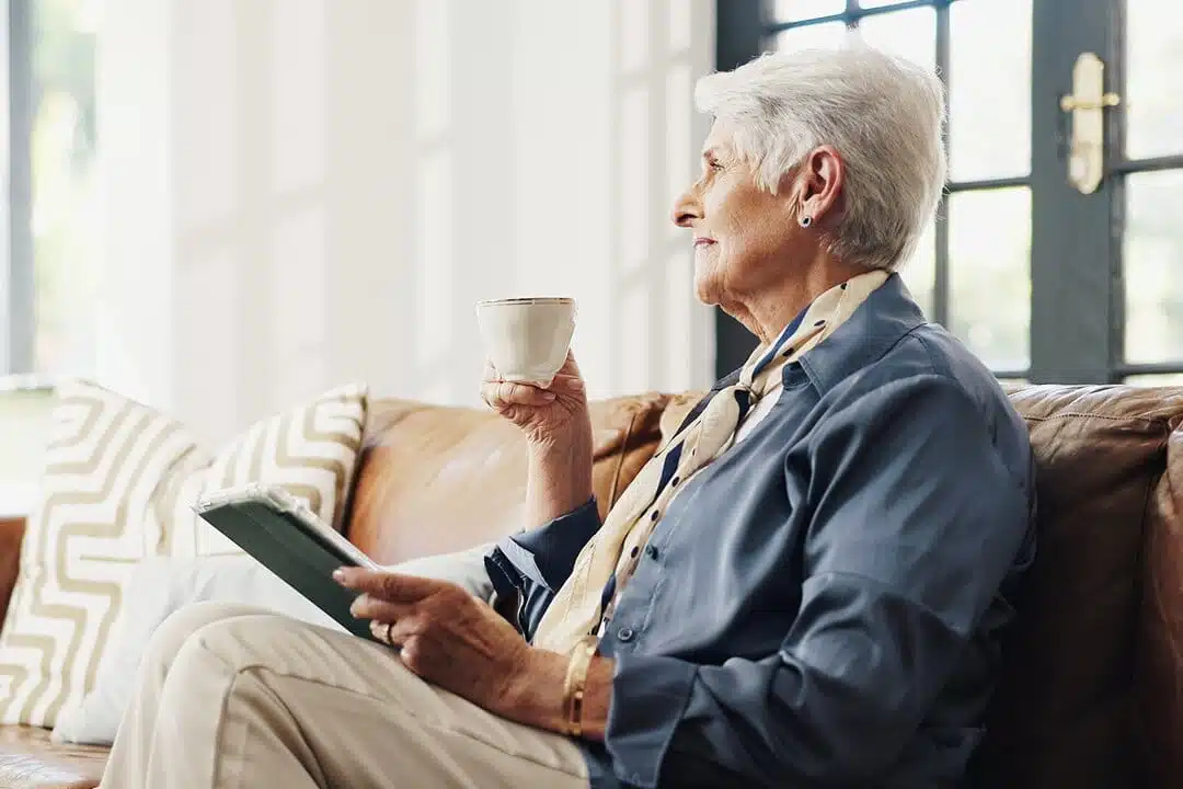 Une élégante dame d'un certain âge, aux cheveux blancs coupés courts, est assise sur un canapé en cuir, une tasse de thé et une tablette à la main, le regard tourné vers la fenêtre baignée de soleil