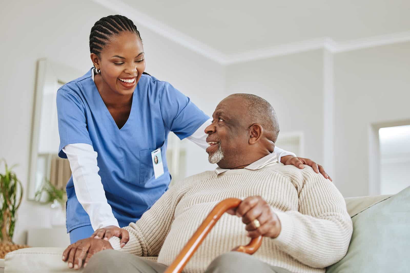 A smiling female healthcare worker in blue scrubs with her hand on the shoulder of an elderly man seated on a sofa. The man is holding a wooden cane and looking up at her with a smile.