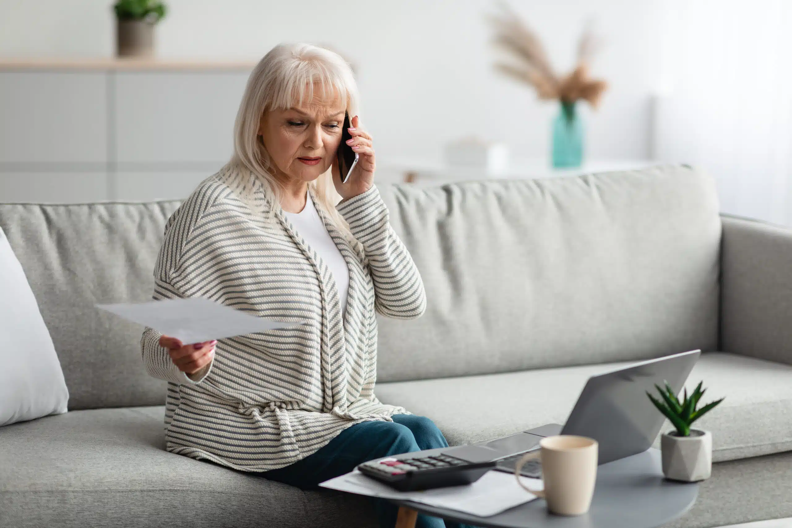 Older woman sitting while on the phone and confused about a paper she is holding while looking at her laptop